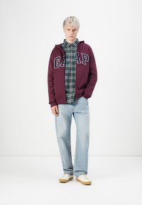 Jong persoon in een bordeauxrode GAP-hoodie over een groen geruit shirt, lichte spijkerbroek en witte sneakers, staand tegen een eenvoudige witte achtergrond.