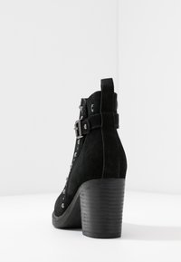 Botín de ante negro hasta el tobillo con un tacón grueso, que cuenta con una cremallera lateral, detalle de hebilla y tachuelas metálicas en la parte delantera.