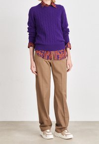 Lila kabelstickad tröja över en mönstrad orange och lila skjorta med krage, kombinerad med beige vida byxor och krämfärgade sneakers.