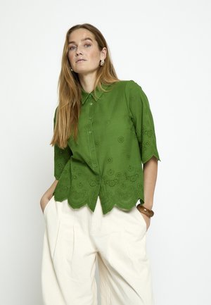 Frau mit langen hellbraunen Haaren, die eine grüne Bluse mit Knöpfen, Ösen-Stickereien und gezackten Kanten trägt, kombiniert mit locker sitzenden weißen Hosen.