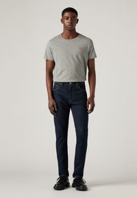 Joven de pie con una camiseta gris de Levi's, jeans azul oscuro y zapatos negros de cordones, contra un fondo claro y liso.