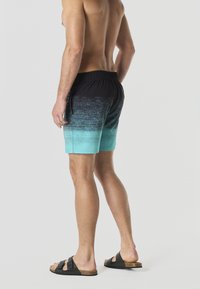 Modello maschile in piedi di profilo, indossa pantaloncini da bagno a gradiente nero e verde acqua e sandali neri a doppio cinturino su uno sfondo grigio uniforme.