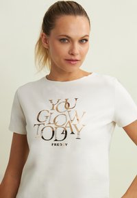 Maglietta bianca in cotone con una texture dorata e nera stampata con lo slogan "YOU GLOW TODAY" e il nome del marchio "FREDDY" in caratteri in grassetto.