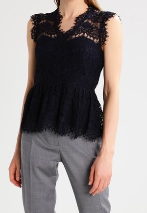 Sort blondebluse uden ærmer med peplumkant båret med lysegrå skræddersyede bukser på en stående person.