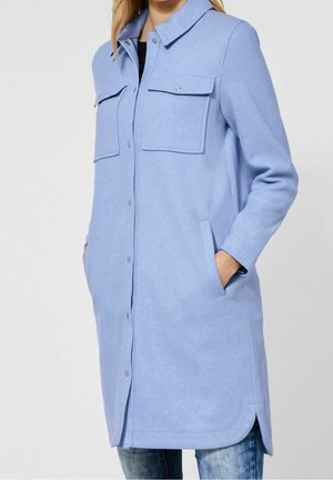 Žena v dlouhém světle modrém kabátu s předními druky, dvěma náprsními kapsami s klopy, bočními kapsami a zaobleným lemem přes džíny.