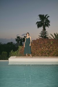 Femme en robe vert foncé debout près d'une piscine au crépuscule avec des palmiers et des arbustes en arrière-plan.