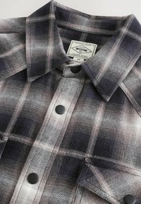 Ruudullinen flanellipaita mustassa, harmaassa ja valkoisessa, jossa on napitettava etuosa, suuri kaulus ja mustat napit; tarrassa lukee "Premium Grade" ja koko "M."