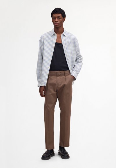 Homme debout portant un pantalon marron, un débardeur noir, une chemise à carreaux bleu clair ouverte et des chaussures en cuir noir, sur fond blanc.