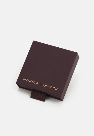 Ruskea suorakulmainen laatikko, jossa on teksturoitu pinta. Siinä on kultainen logo, jossa lukee "MONICA VINADER" ja vetotaba avaamista varten.