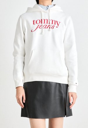 Persoon draagt een witte hoodie met rode "tommy jeans" tekst, daaronder een lichtblauw overhemd met kraag, en een zwarte leren rok met een split aan de voorkant.