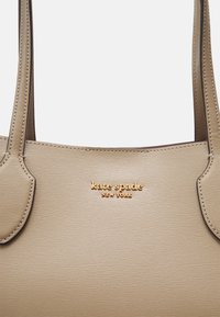 Beige skinnveske med teksturert overflate, med doble håndtak og gullfolie "kate spade NEW YORK" logo foran.