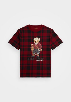 T-shirt en coton à carreaux rouges et noirs avec un graphique d'ours vêtu d'une tenue en flanelle rouge, accompagné d'un texte imprimé sous l'illustration.