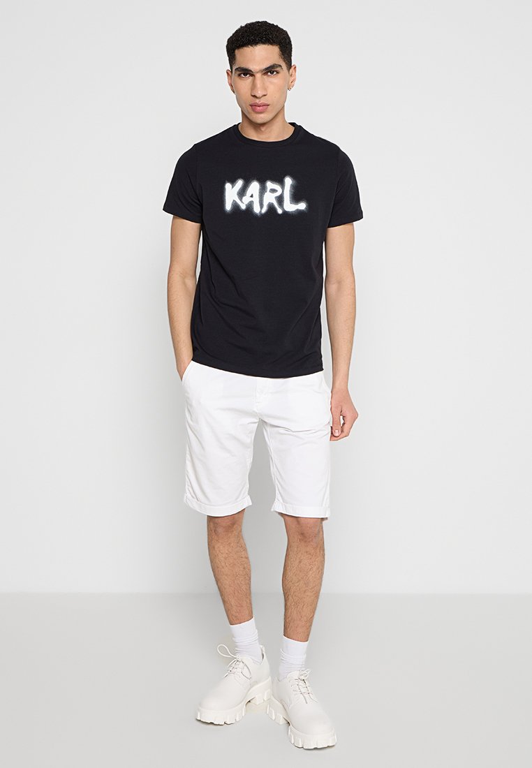 T-shirt noir avec un graphique blanc "KARL", porté avec un short blanc et des baskets blanches imposantes. Le modèle a des manches courtes et un col rond.