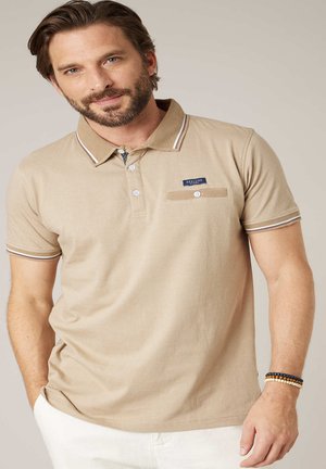 Homme barbu portant un polo beige avec des boutons blancs, une petite poche poitrine, et des bracelets au poignet droit, posant devant un fond uni.