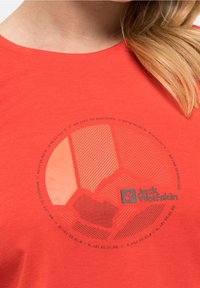 Detailní záběr na červenou košili s kruhovým geometrickým vzorem a logem "Jack Wolfskin" na hrudi, kterou nosí osoba s blond vlasy.