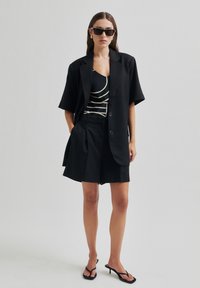 Černý přizpůsobený blejzr s volným střihem, doplněný černými šortkami s vysokým pasem. Modelka má na sobě pruhované tričko a černé sandály na podpatku.