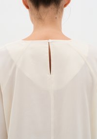 Blusa crema con una delicata texture e un'apertura a oblò sul retro, chiusa da un singolo bottone. Design semplice e fluido.