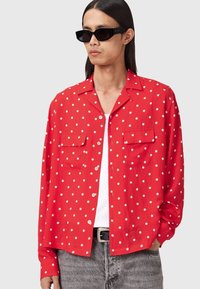 Rød kortærmet skjorte med hvide prikker, to brystlommer og en knapstolpe. Båret over en hvid tanktop, parret med mørke jeans.