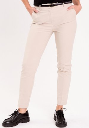 Persona che indossa pantaloni beige slim-fit, cintura nera e scarpe nere chunky stringate, in piedi con le mani in tasca.