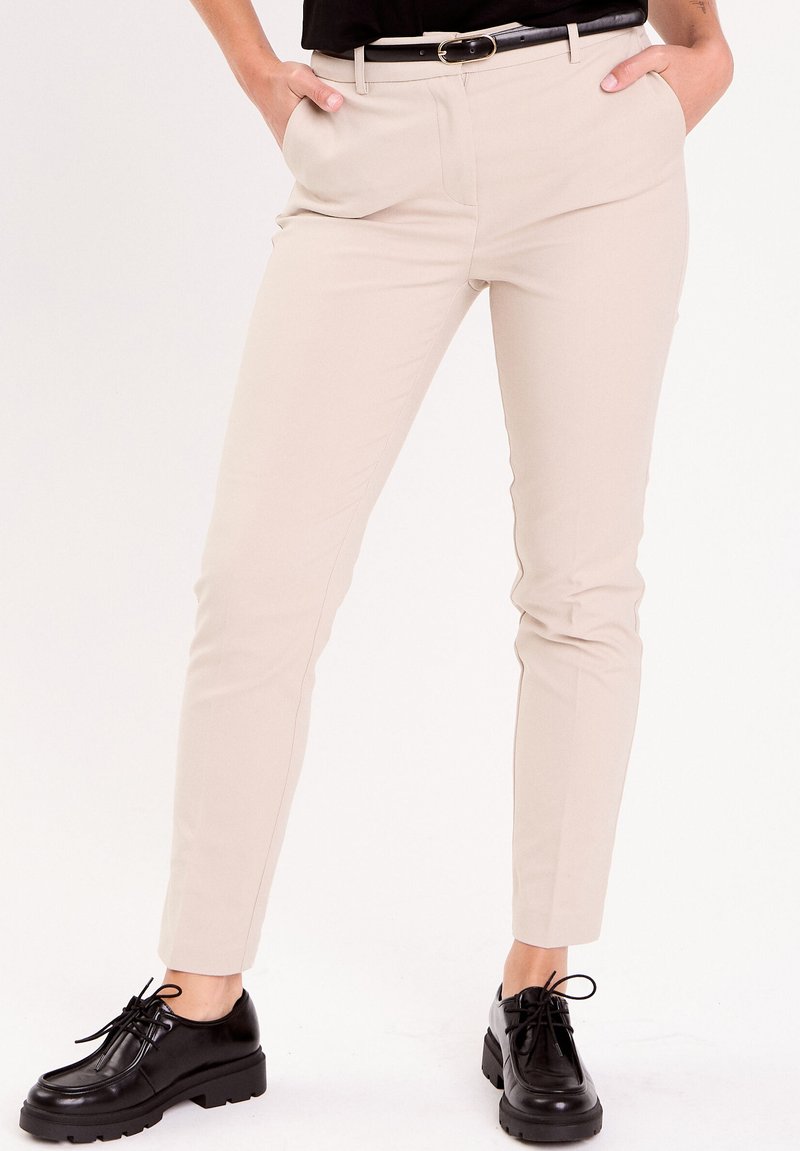 Persoon draagt beige slim-fit broek, zwarte riem en zwarte veterchunky schoenen, staand met handen in de zakken.