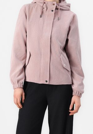 Femme portant une veste à capuche en velours côtelé rose poussiéreux avec des boutons-pression, associée à un pantalon noir, sur fond blanc uni.