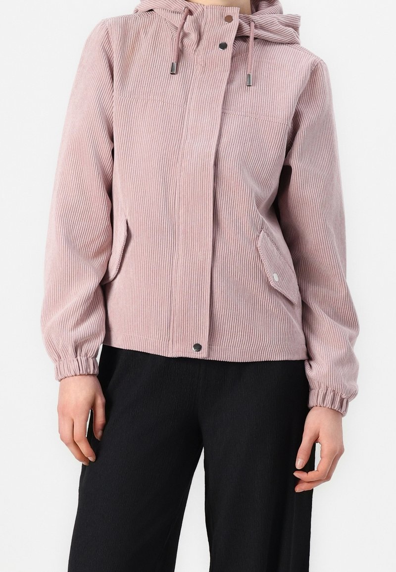 Femme portant une veste à capuche en velours côtelé rose poussiéreux avec des boutons-pression, associée à un pantalon noir, sur fond blanc uni.