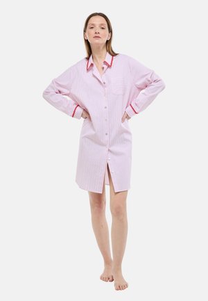Femme debout pieds nus portant une robe-chemise rose clair à manches longues avec un liseré rouge au col et aux poignets, mains sur les hanches, expression neutre.