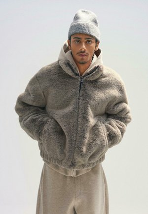 Faux fur zip-up jakke i lys grå, med oversized silhuet, høj krave, elastisk kant og store forlommer. Sammen med en grå hue.