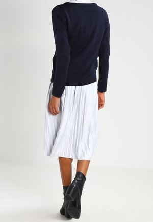 Femme portant un pull bleu marine, une jupe midi plissée blanche et des bottines noires, debout dos tourné sur un fond uni.