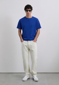 Hombre con gafas de sol negras, camiseta azul, pantalones crema y zapatillas blancas, de pie con las manos en los bolsillos sobre un suelo gris frente a una pared blanca.