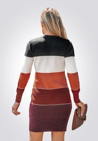 Gebreide jurk van wol met zwarte, witte, oranje en bordeauxrode horizontale strepen. Ribgebreide mouwen en zoom. De textuur lijkt zacht en behaaglijk.