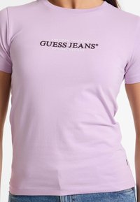 Šviesiai purpurinė medvilninė marškinėliai su juodu „GUESS JEANS“ logotipu centre, trumpomis rankovėmis ir prigludusia forma.
