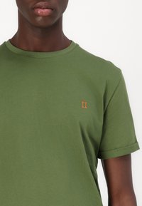 T-shirt verde de algodão com mangas enroladas e um pequeno bordado laranja "II" no peito. Textura suave e decote redondo clássico.