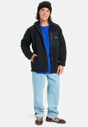 Czarna polarowa kurtka z zamkiem i kieszenią, niebieska koszulka, jasnoniebieskie dżinsy oraz brązowe sneakersy z białymi paskami. Model nosi czarną czapkę beanie.