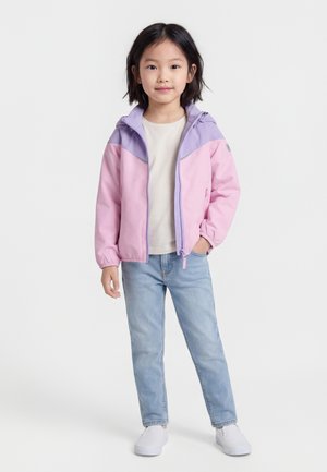 Κορίτσι στέκεται με το ένα χέρι στην τσέπη φορώντας ροζ και λιλά μπουφάν, λευκό πουκάμισο, ανοιχτό μπλε τζιν και λευκά παπούτσια slip-on σε λευκό φόντο.