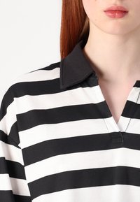 Pruhovaná polo košile v černobílé barvě s černým límečkem, obsahující klopu a měkkou texturu látky. Jsou viditelné pečlivě ušité detaily.