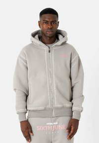 Grijze zip-up hoodie met decoratieve witte stiksels, kangoeroe-instapk pockets en een capuchon. Kenmerkt het logo "SIXTH JUNE" in het roze op de borst.