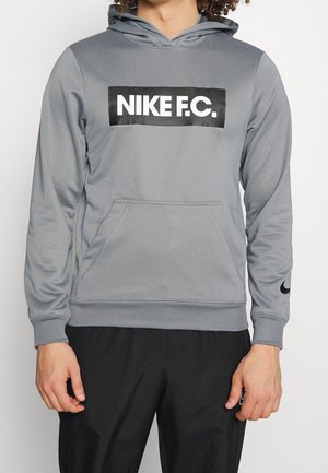 Osoba nosząca szarą bluzę z kapturem z napisem "NIKE F.C." na piersi oraz czarnym logo Nike swoosh na mankiecie lewego rękawa.