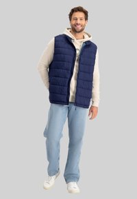 Marineblauer Puffer-Weste mit gestepptem Design, ausgestattet mit einem Vorderreißverschluss und Kapuze. Getragen über einem hellgrauen Hoodie, kombiniert mit hellblauen Jeans und weißen Sneakers.