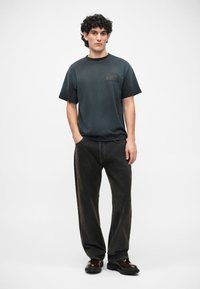 Dunkelgraues Baumwoll-T-Shirt mit kurzen Ärmeln und einem Logo auf der Brust, kombiniert mit locker sitzenden schwarzen Jeanshosen und klobigen schwarzen Schuhen.