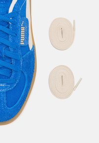 Delvis vy av en blå Puma-sneaker med mockadetaljer och två vita snurrade skosnören mot en vit bakgrund.