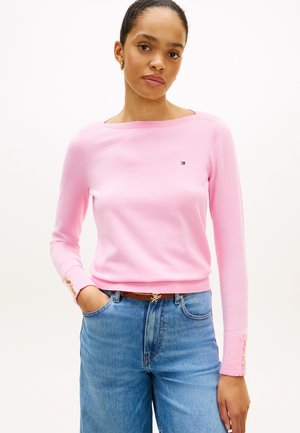 Frau trägt einen hellrosa Pullover mit langen Ärmeln und goldenen Knöpfen an den Manschetten sowie eine hoch taillierte blaue Jeans und steht mit einer Hand in der Tasche.