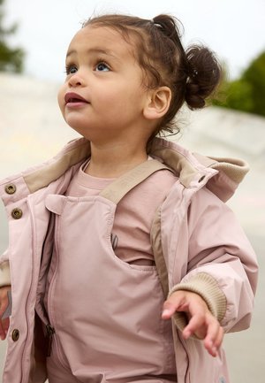 Hellrosa isolierte Jacke mit beiger Innenauskleidung, kombiniert mit einer passenden Latzhose. Weiche Textur, Druckknopfverschlüsse und sichtbare Schulterträger.