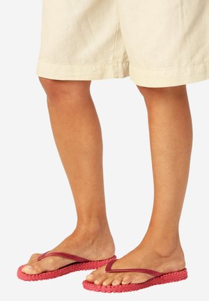 Persona che indossa pantaloncini color crema fino al ginocchio e infradito rosse con suole strutturate, in piedi su sfondo bianco.