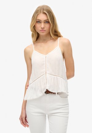 Giovane donna con capelli biondi lunghi indossa una canottiera bianca con dettagli in pizzo e pantaloni bianchi, posando davanti a uno sfondo neutro.