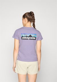 T-shirt en coton violet clair avec un logo noir Patagonia et un graphisme de montagne, accompagné de rayures colorées dans le dos. Manches courtes, coupe décontractée.