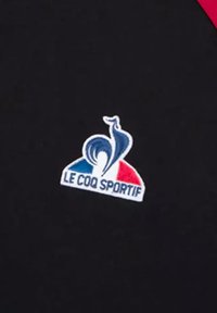 Zwarte shirt met een gekleurde logo van "Le Coq Sportif" met een gestileerde hen, met blauwe, witte en rode accenten in borduurwerk.