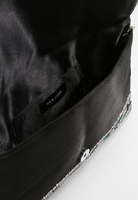 Interior de un bolso de satén negro, con una textura suave, herrajes plateados y una etiqueta de "NEW LOOK". Cierre con un detalle de botón circular.