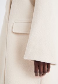 Manteau en laine couleur crème, présentant une texture douce, des manches larges et une poche poitrine unique, avec un gros plan d'une main reposant sur le tissu.