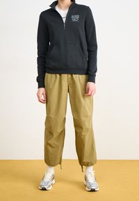 Svart sweatshirth med dragkedja och framficka, kombinerad med löst sittande olivgröna byxor och gråvita sneakers med svarta detaljer.